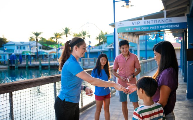 A SeaWorld guide in a blue polo hands something to a smiling mother as her family of four gathers on a boardwalk near a VIP Entrance sign reading We love our Pass Members