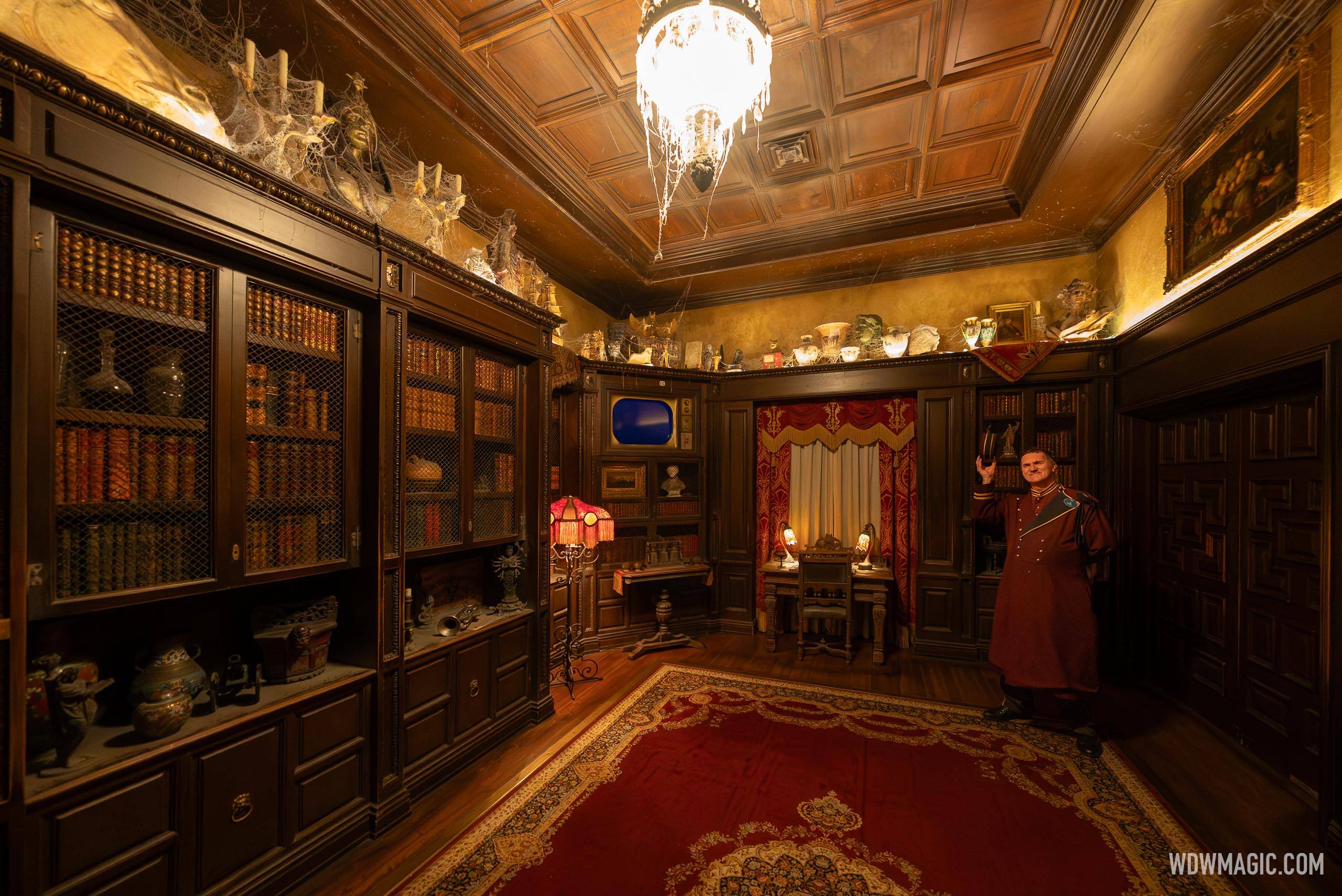 A dim, cobwebbed library with wood bookshelves, a vintage television set in the back wall, a red rug, and a Cast Member in a burgundy bellhop uniform tipping his cap