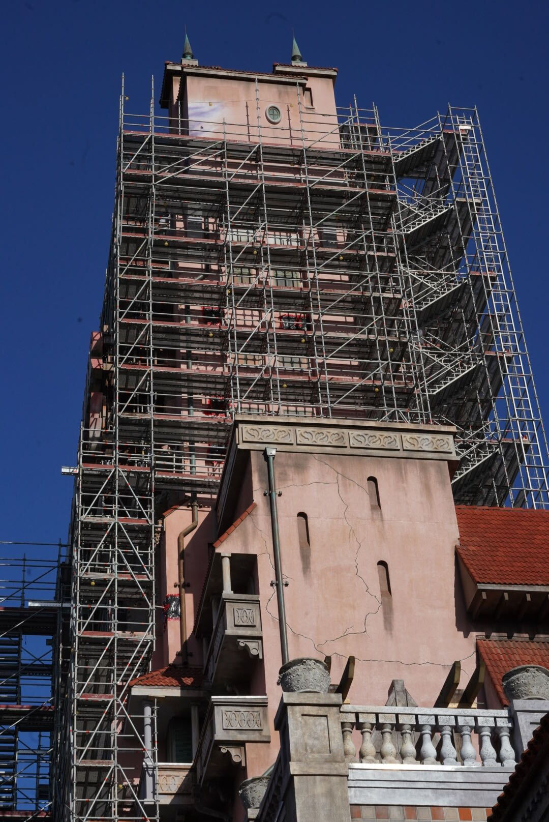Looking up at the scaffolded Hollywood Tower Hotel against a deep blue sky, with a pale purple streak near the top beside a round window and painted cracks running down a lower pink wall
