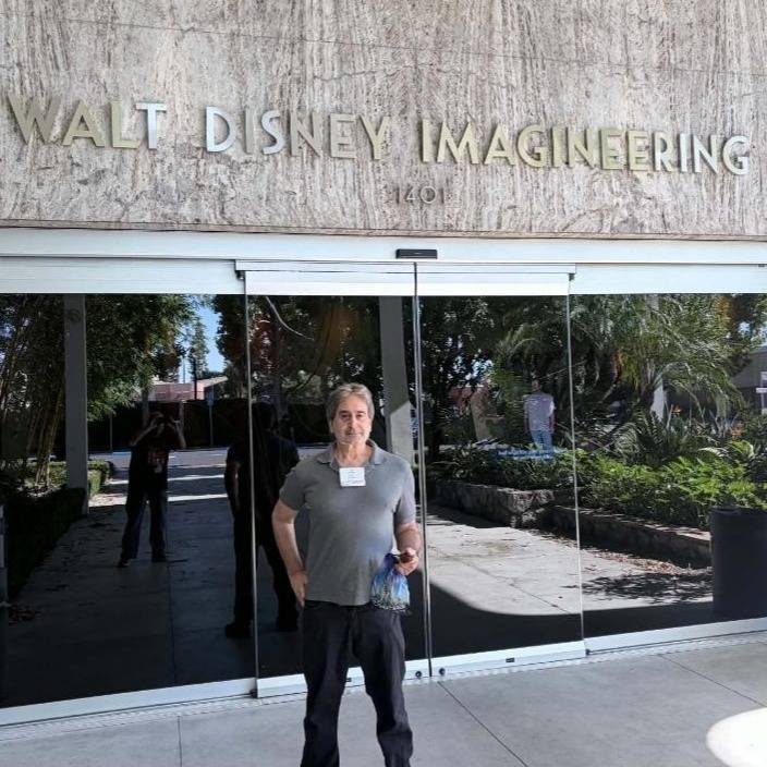 A man in a gray polo shirt and visitor badge stands in front of glass doors beneath a stone wall lettered Walt Disney Imagineering 1401