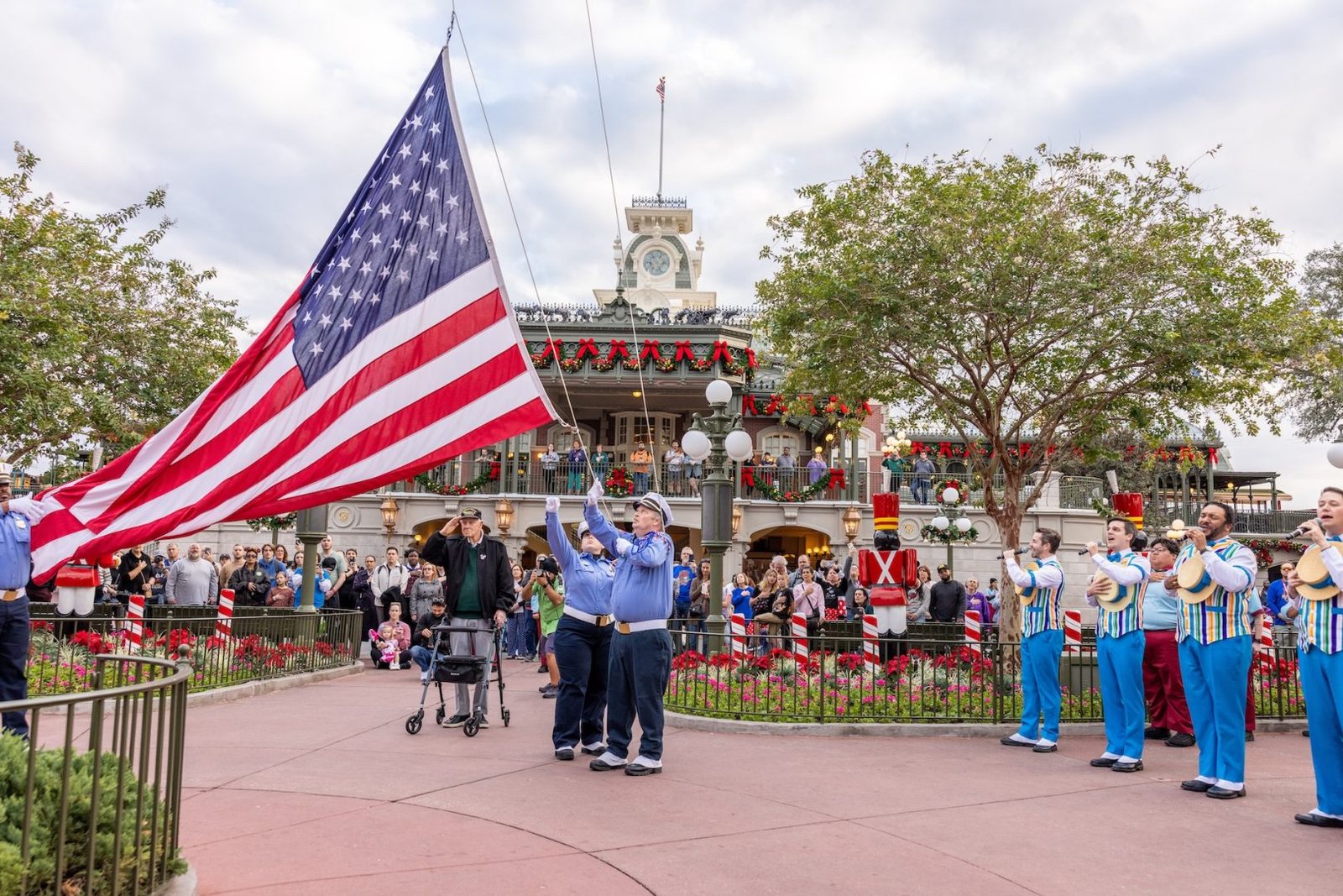 Cast members lower a large American flag in Town Square at Magic Kingdom as an older man standing beside a walker salutes and singers in striped vests perform, with holiday garlands on the train station behind