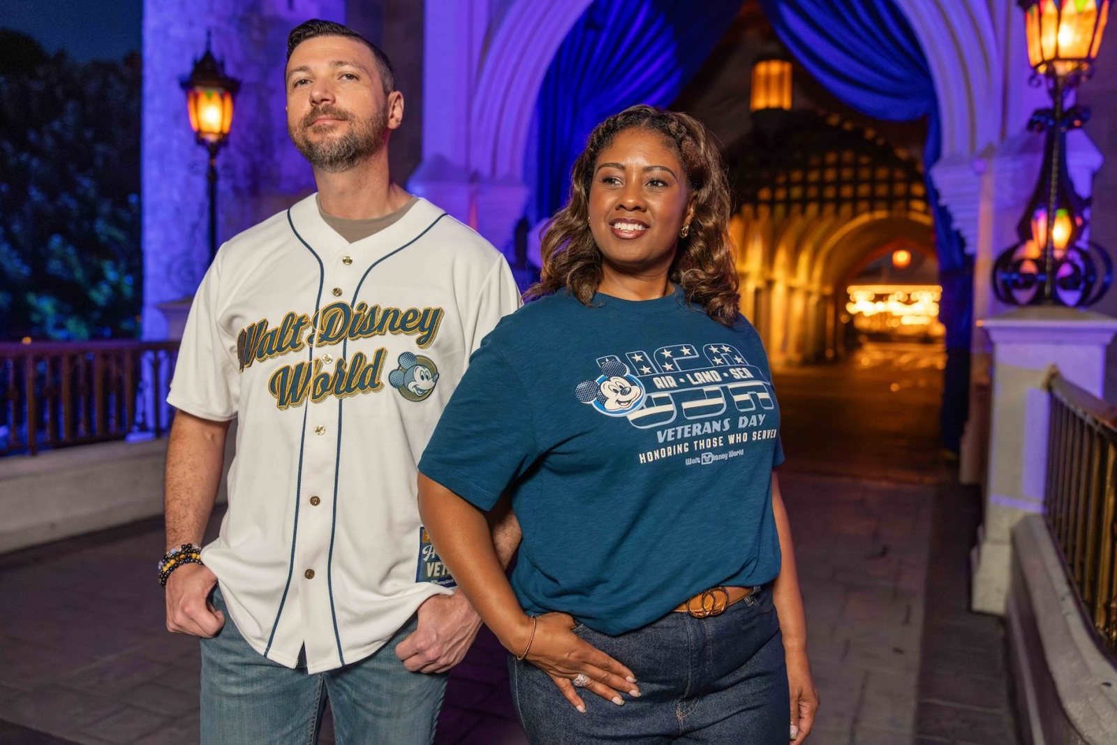 A man in a white Walt Disney World baseball jersey and a woman in a blue USA Air Land Sea Veterans Day T-shirt stand under a lantern-lit archway at night