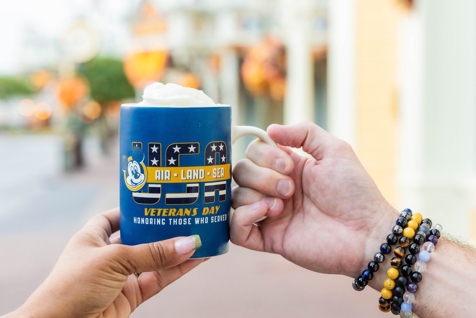 Two hands hold a blue Veterans Day mug topped with whipped cream, showing USA Air Land Sea art, with a blurred Main Street in the background