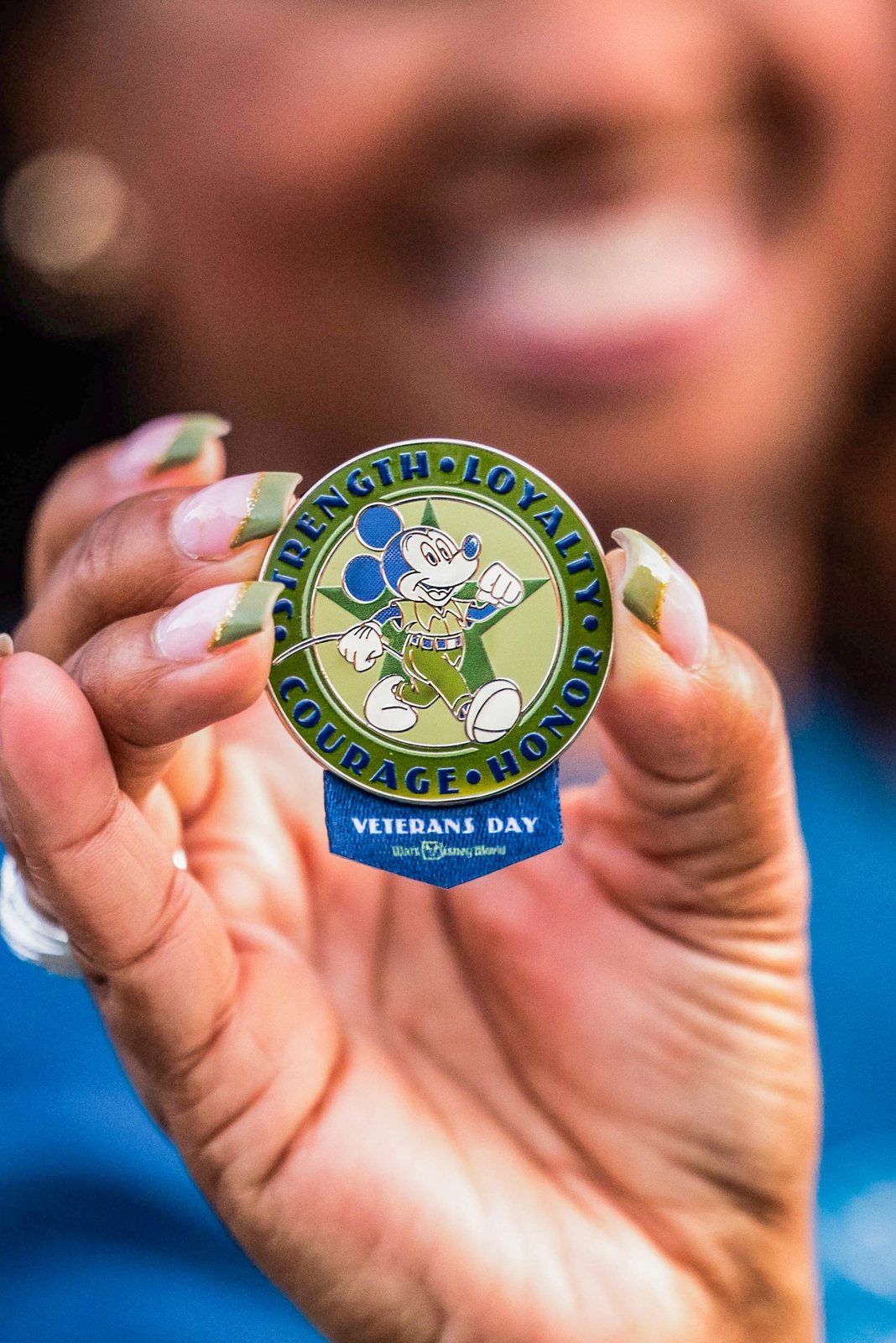 A hand holds up a round green and blue pin showing Mickey Mouse in army green, ringed by the words Strength Loyalty Courage Honor, with a Veterans Day banner at the bottom