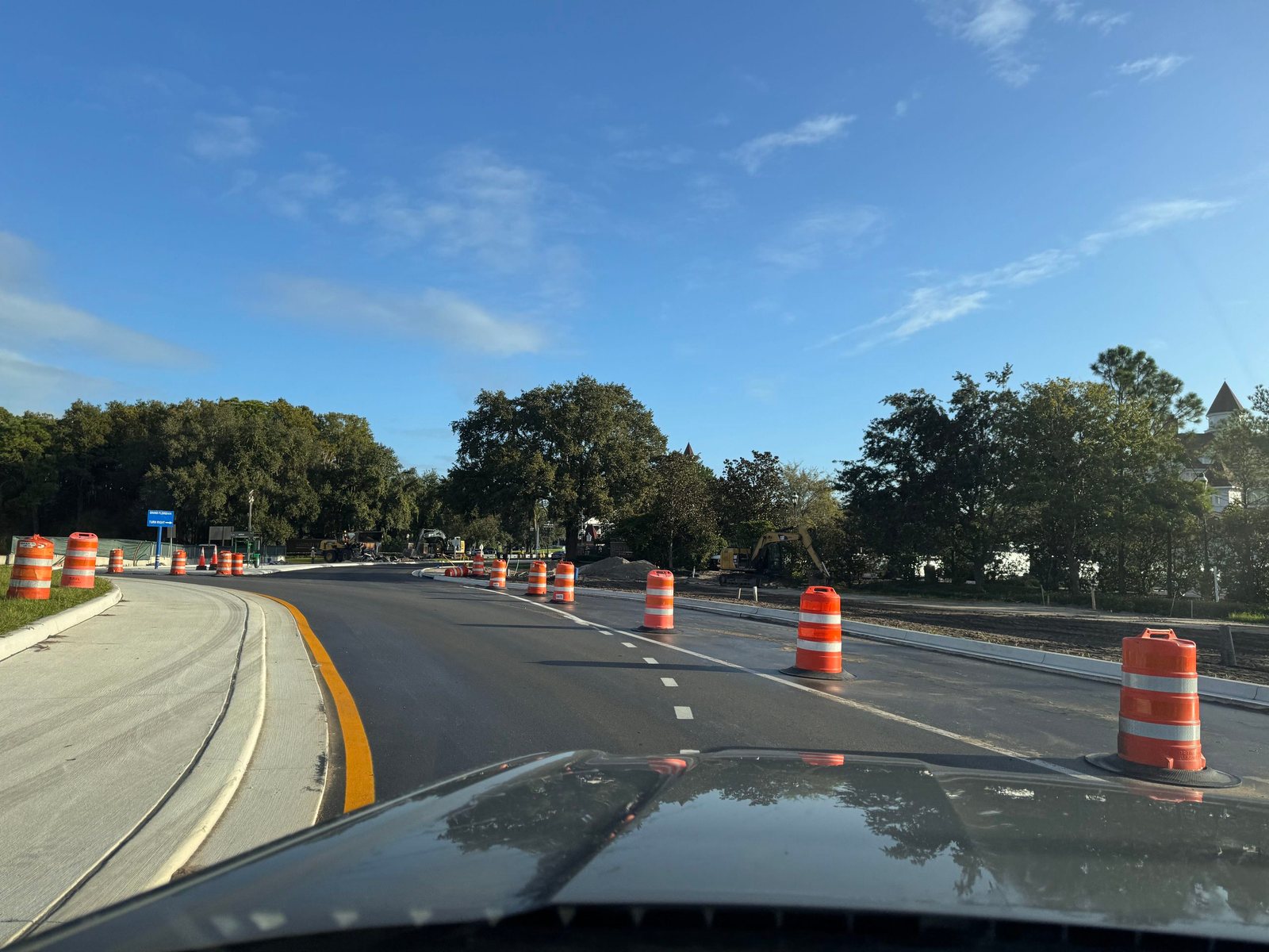 View through a car windshield along a curving newly paved road lined with orange and white barrels, with a blue Grand Floridian Turn Right sign at left, excavators beside the road, and a Grand Floridian turret visible above the trees at right