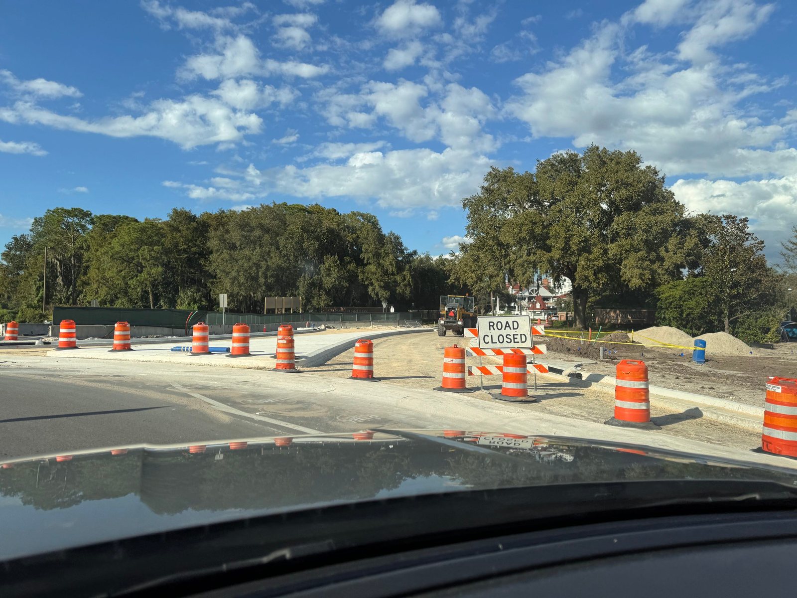 View through a car windshield at a roundabout exit blocked by a Road Closed sign and orange barrels, with an unpaved road, a front loader and sand piles beyond, and the Grand Floridian entrance sign and a white Victorian building behind trees