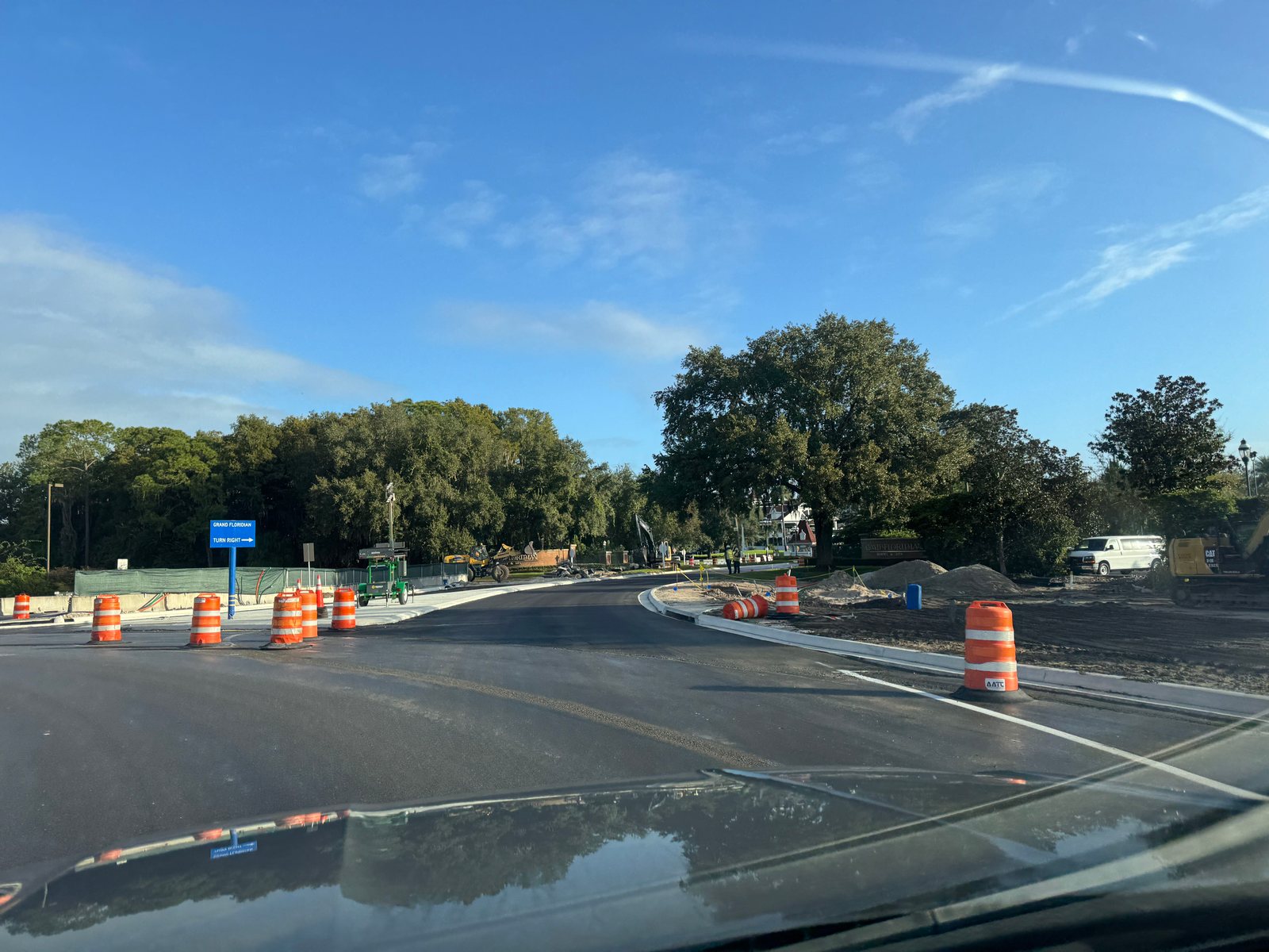 View through a car windshield of a freshly paved road leaving a roundabout, lined with orange and white construction barrels, with a blue sign reading Grand Floridian Turn Right at left and construction equipment and the Grand Floridian entrance sign in the distance