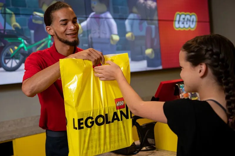 A smiling Legoland team member in a red polo shirt hands a yellow LEGOLAND shopping bag across a counter to a girl, with a large LEGO screen display behind him