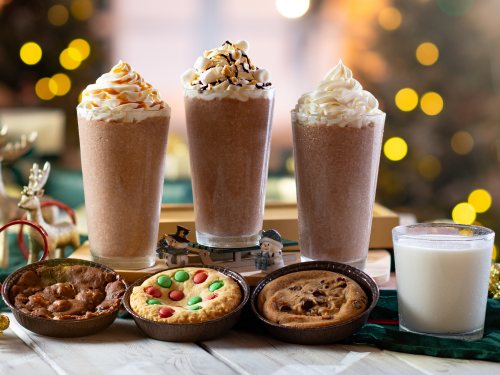 Three tall frozen cocoa drinks topped with whipped cream and drizzle, a glass of milk, and three cookies in small tins, one topped with candy-coated chocolates, on a wooden table in front of blurred Christmas tree lights