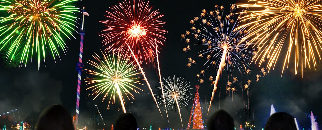 Red, green, white and gold fireworks bursting over SeaWorld Orlando at night above a lit Christmas tree and the striped Sky Tower, with guests silhouetted in the foreground