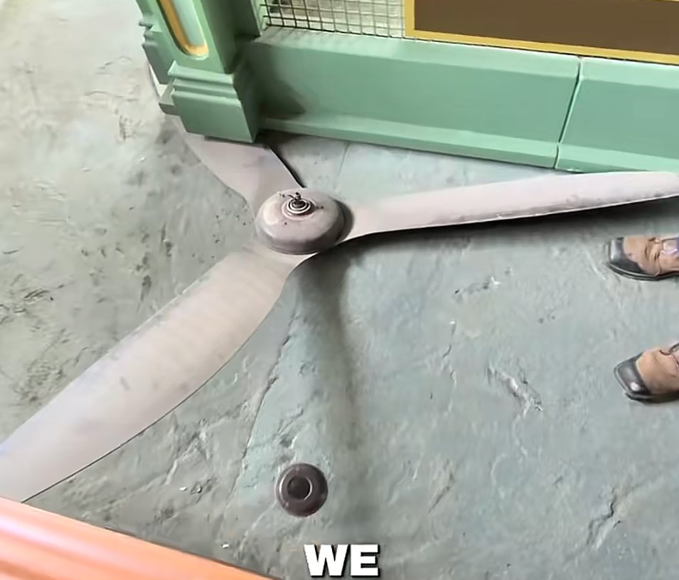 A large three-bladed ceiling fan lying on a grey concrete queue floor against a mint green wall, one blade wedged under the corner of the wall, with a guest's shoes visible at the right edge and the caption word WE burned into the bottom of the frame