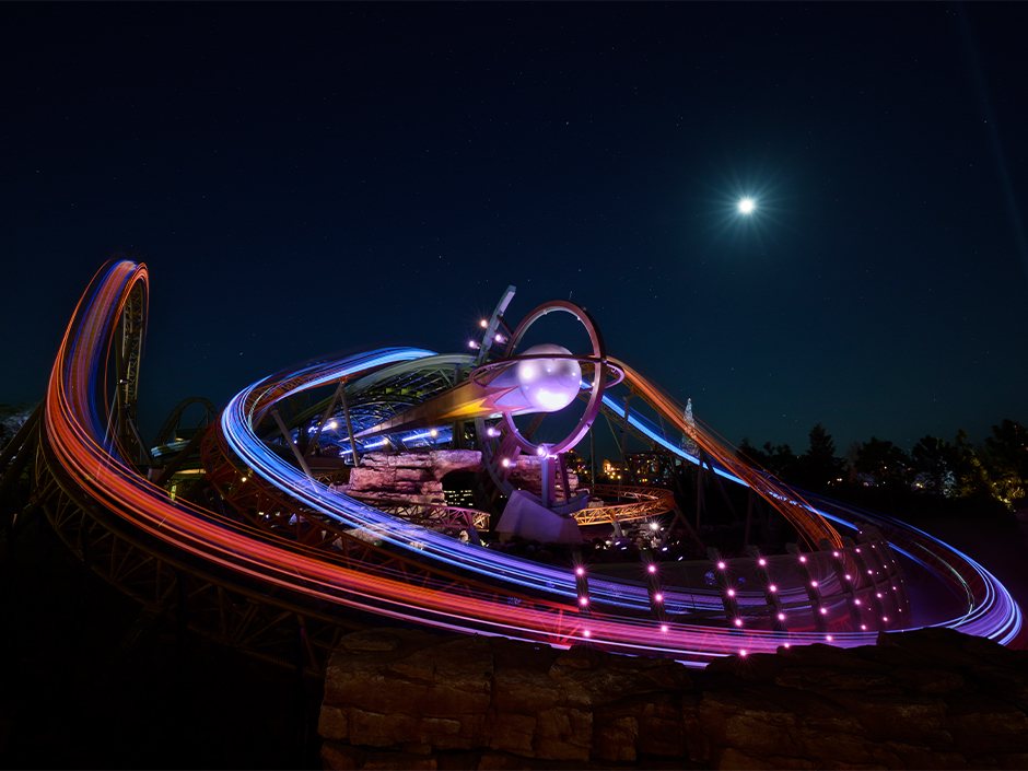 Long exposure of Stardust Racers at night, red and blue light trails looping around the globe and ring centerpiece under a bright moon