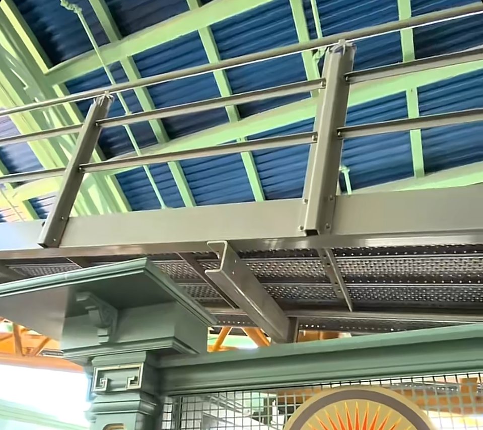 Looking up at a grey metal work ramp with railings and perforated decking beneath a blue corrugated roof and pale green steel frame, above a green queue wall and a sunburst emblem