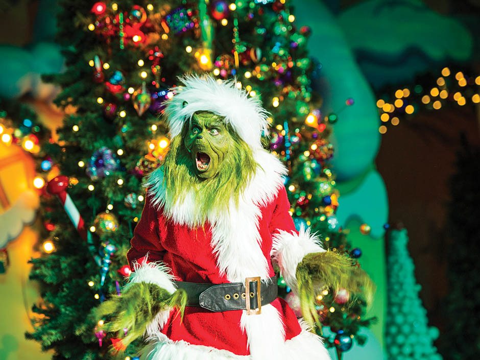The Grinch in a red and white Santa suit with a black belt, mouth wide open, in front of a decorated Christmas tree