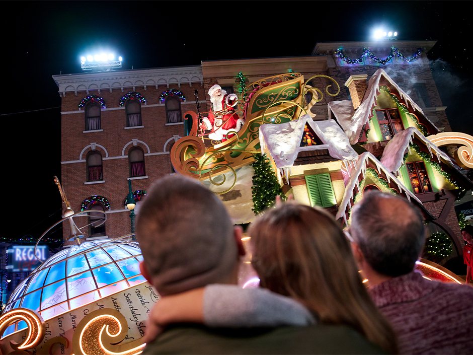 Guests seen from behind watch Santa Claus wave from a gold sleigh float with snowy rooftops during a nighttime parade, with a lit globe float at left