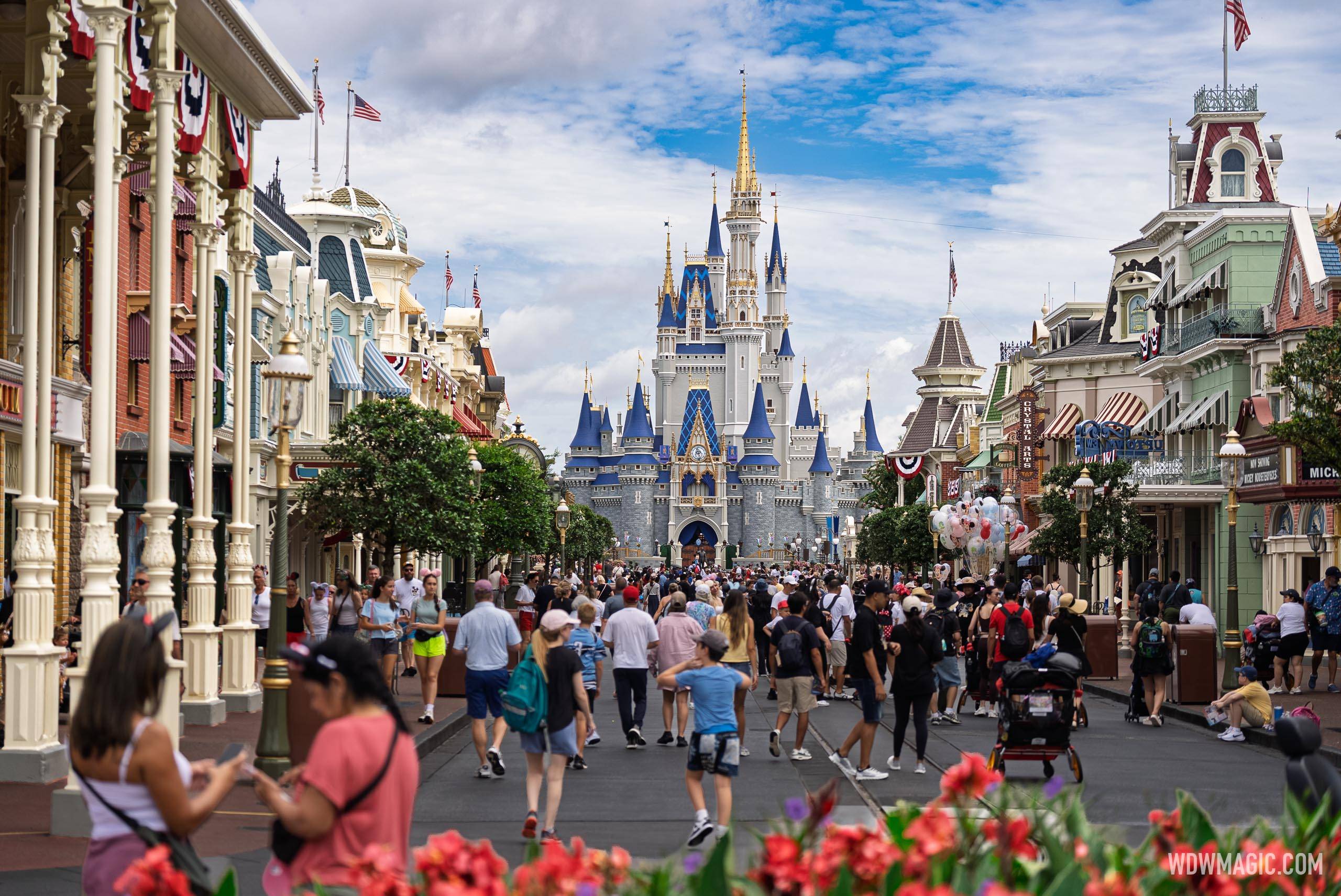 Guests walking down Main Street, U.S.A. at Magic Kingdom toward Cinderella Castle under a blue sky with white clouds, with Victorian storefronts on both sides and red flowers in the foreground