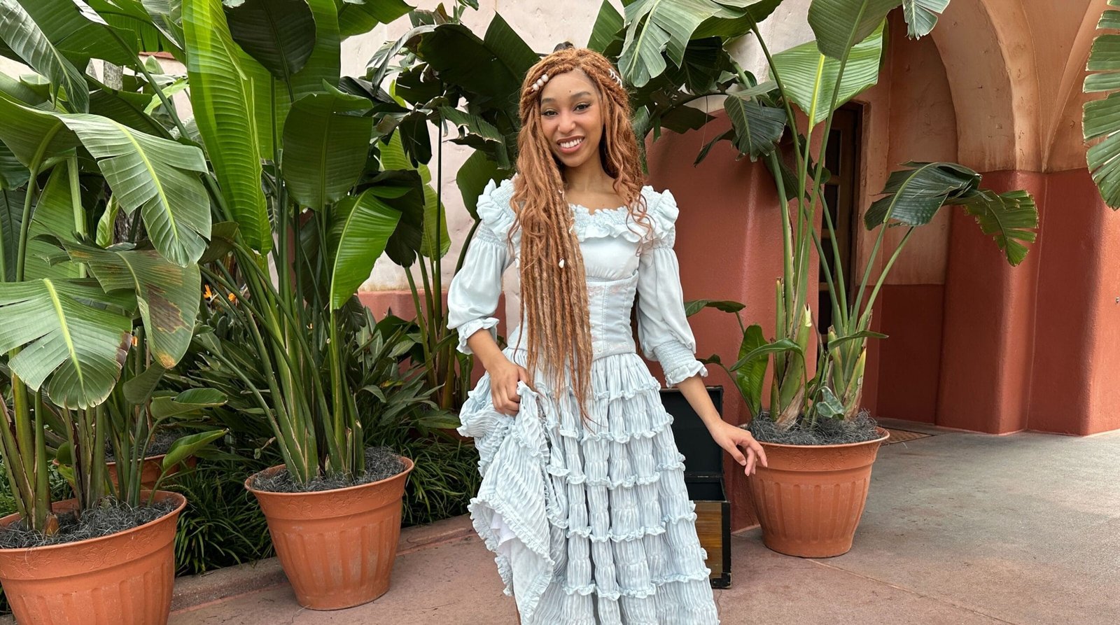 Live-action Ariel in a pale blue tiered ruffle dress, holding her skirt and smiling outdoors in front of large banana plants in terracotta pots and a salmon-colored stucco wall