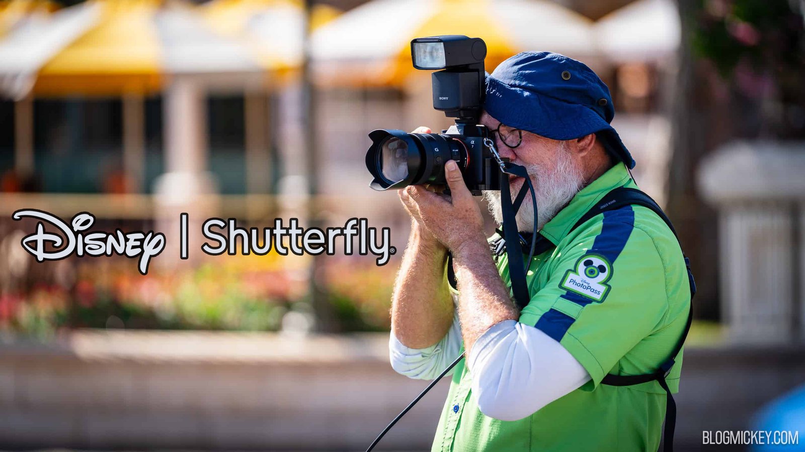 A bearded Disney PhotoPass photographer in a bright green PhotoPass shirt and blue bucket hat shoots with a Sony camera and flash, beside Disney and Shutterfly logos