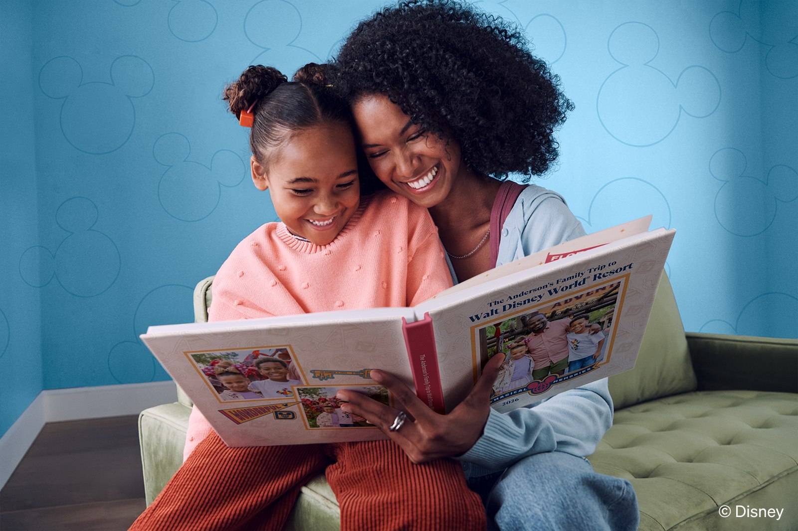 A smiling mother and daughter on a green sofa look through a large photo book titled The Anderson's Family Trip to Walt Disney World Resort, against a blue wall patterned with Mickey Mouse outlines