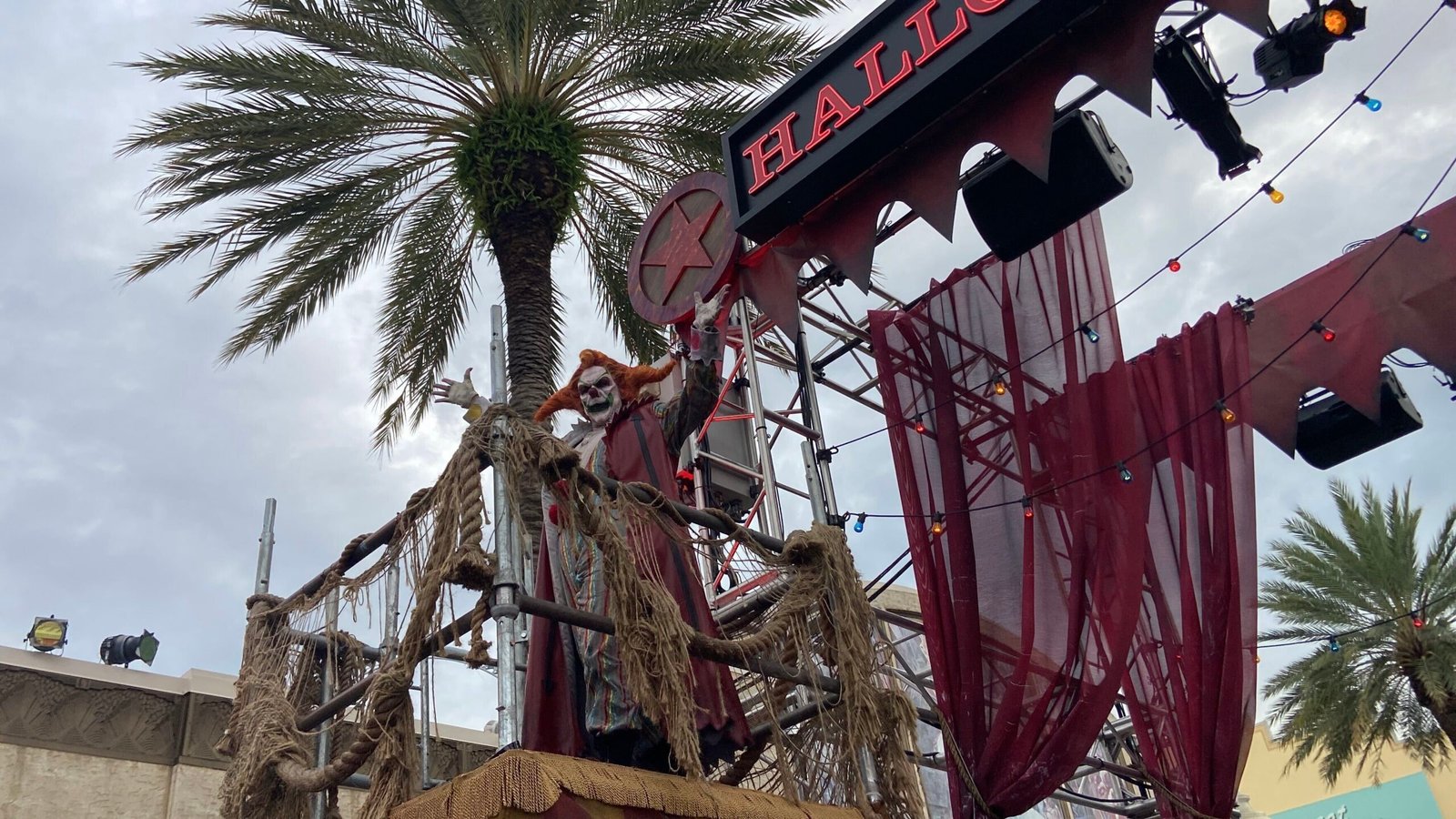 A clown with wild orange hair and skull-like white makeup, both arms raised, standing on a rope-draped platform beside the partly visible red Halloween Horror Nights sign and sheer red drapes under a cloudy sky
