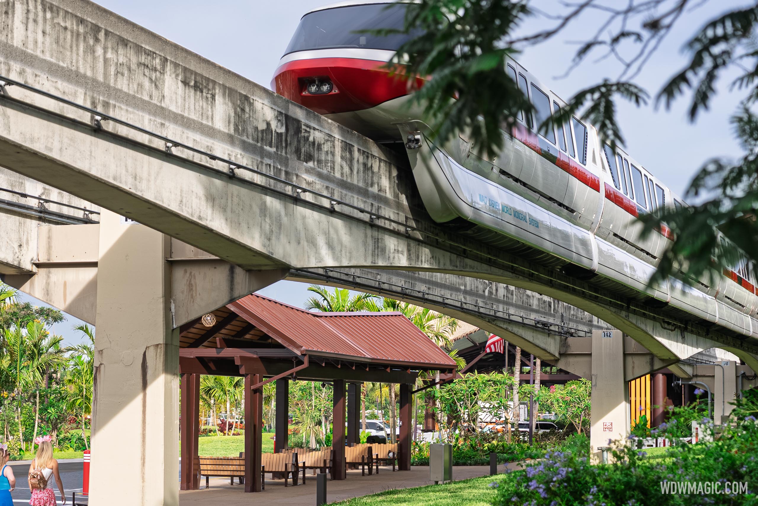 A red-striped monorail passing on its concrete beam above a brown-roofed covered shelter with wooden benches, surrounded by palms and flowering plants
