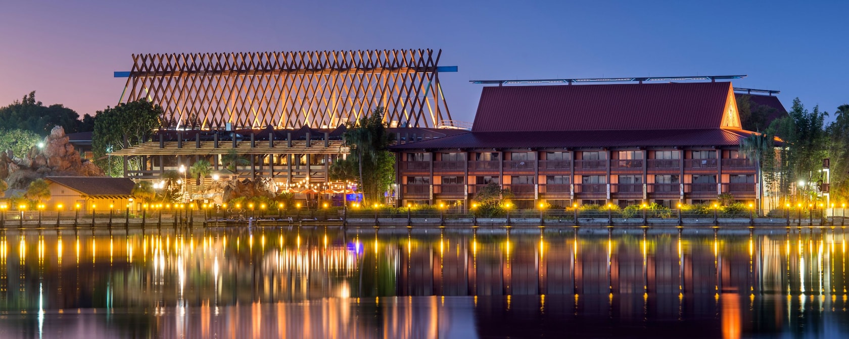 The Great Ceremonial House at Disney's Polynesian Village Resort lit up at dusk, its tall A-frame lattice of roof beams glowing beside a two-story longhouse, with lamps reflected in the water
