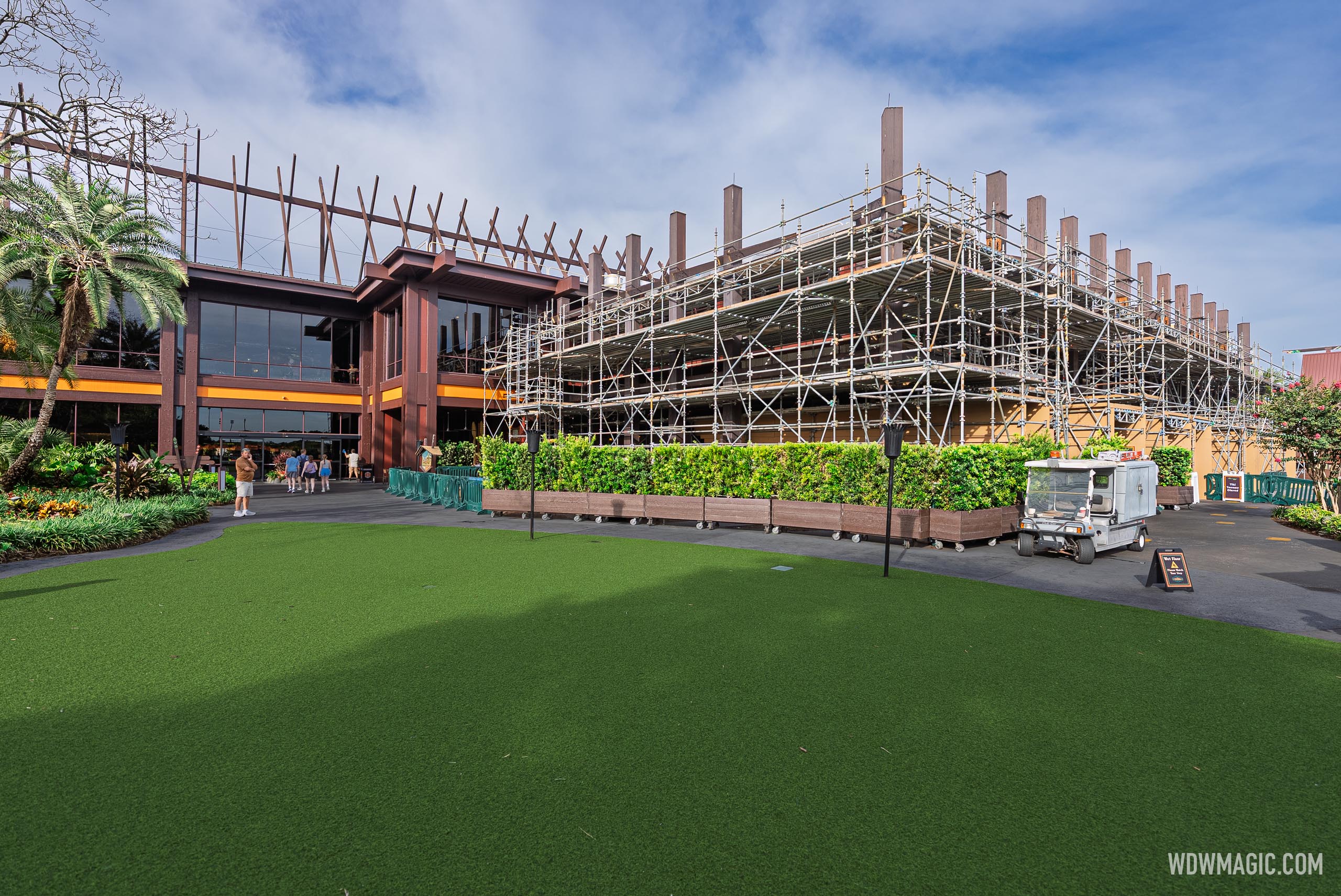 Scaffolding covering one side of the Great Ceremonial House next to its glass entrance, with bare roof beams overhead, a row of planter hedges, a utility cart and an artificial lawn in front