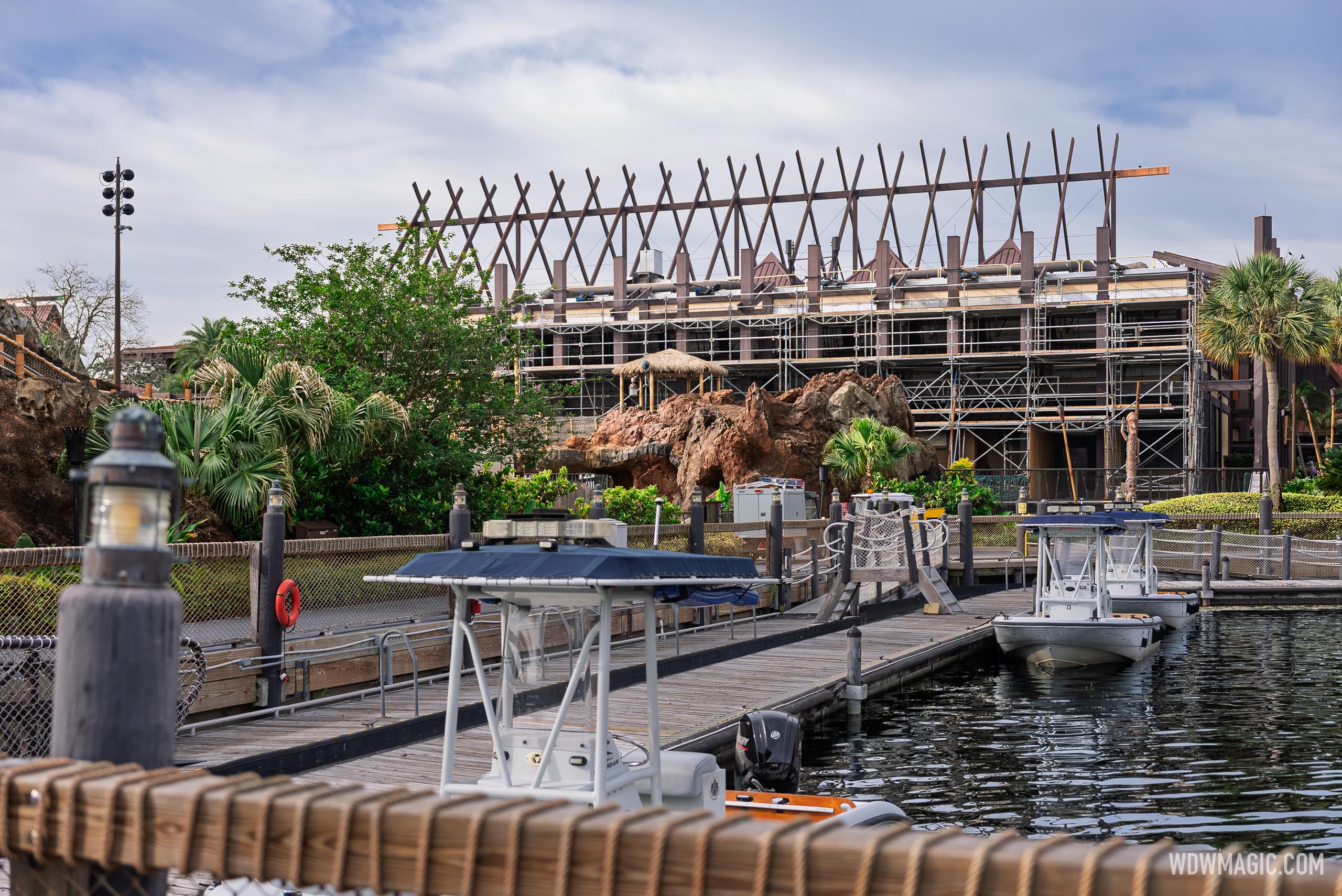 The Great Ceremonial House seen from the Polynesian marina dock, its crossed roof beams rising above a wall of scaffolding, with small boats moored in the foreground