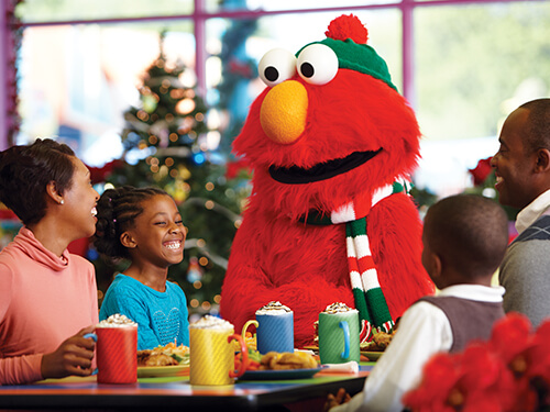 Elmo in a green knit hat and red, white and green striped scarf at a breakfast table with a smiling family, colorful mugs topped with whipped cream in front of them and a decorated Christmas tree behind