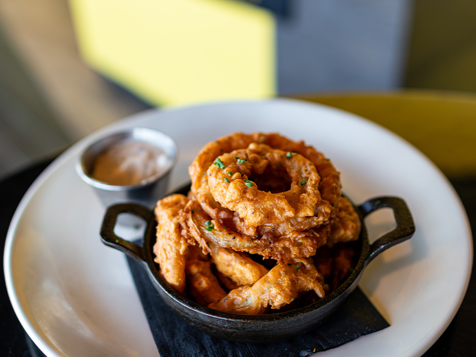 A cast iron skillet piled with battered fried onion rings, one large ring on top, with a small cup of dipping sauce on a white plate