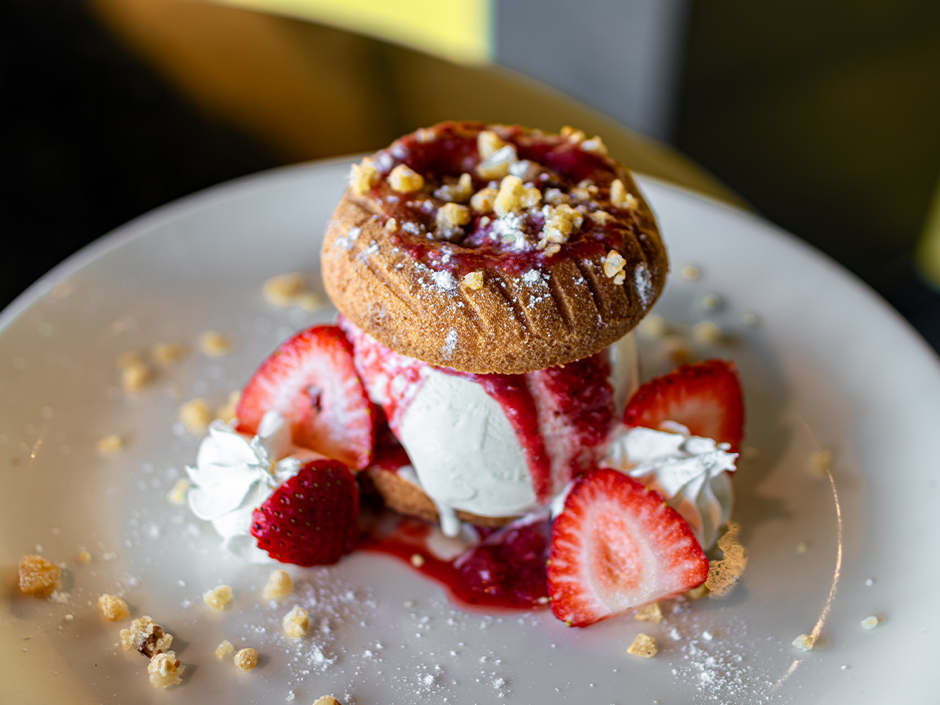 A sugar-dusted cake doughnut set on a scoop of ice cream with red jam, topped with more jam and crumbled brittle, with sliced strawberries and whipped cream on the plate