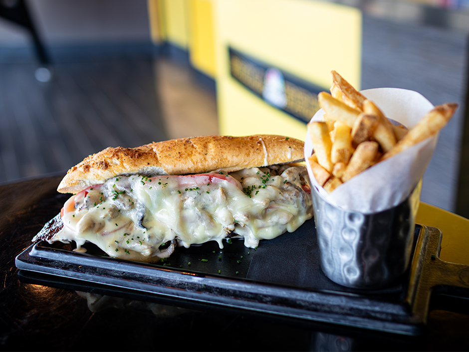 A long sandwich on a pretzel hoagie roll with melted cheese spilling out of it, served on a black board next to a metal cup of fries