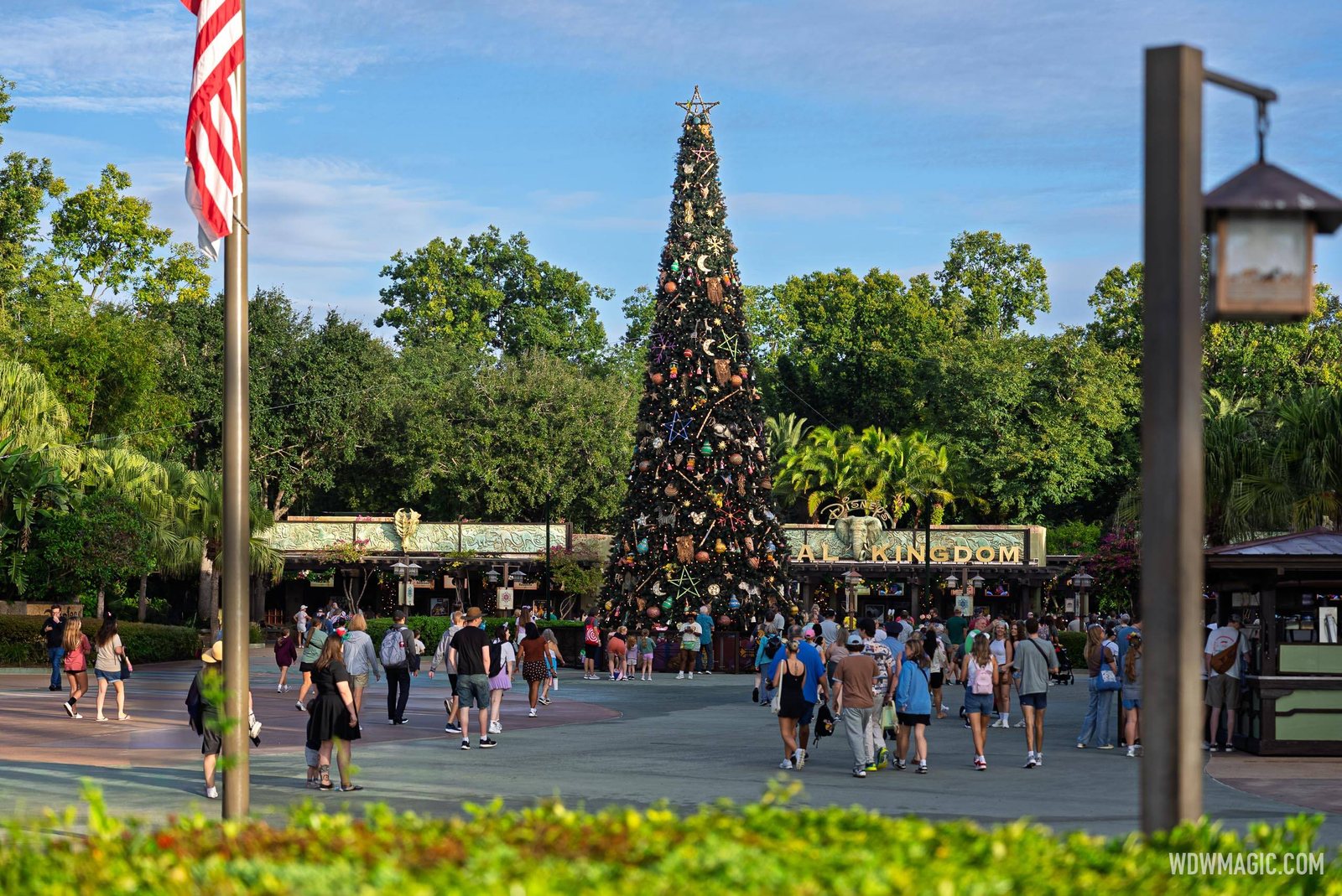 A tall Christmas tree decorated with stars, moons and carved animal ornaments stands in front of the Disney's Animal Kingdom entrance sign, with guests walking across the plaza and an American flag at left
