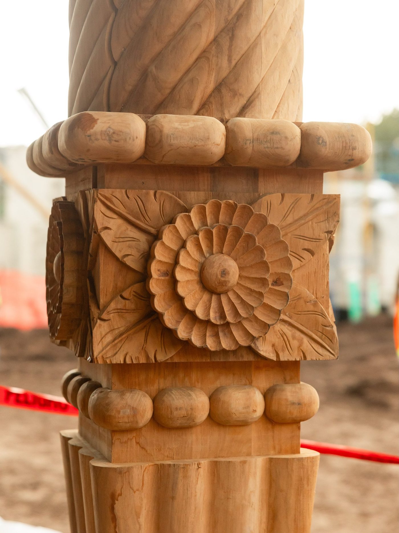 Close-up of a carved wooden column with a large round flower rosette set between carved leaves, a twisted carved shaft above, rounded beading above and below the leaf block, and a fluted base, on a dirt construction site