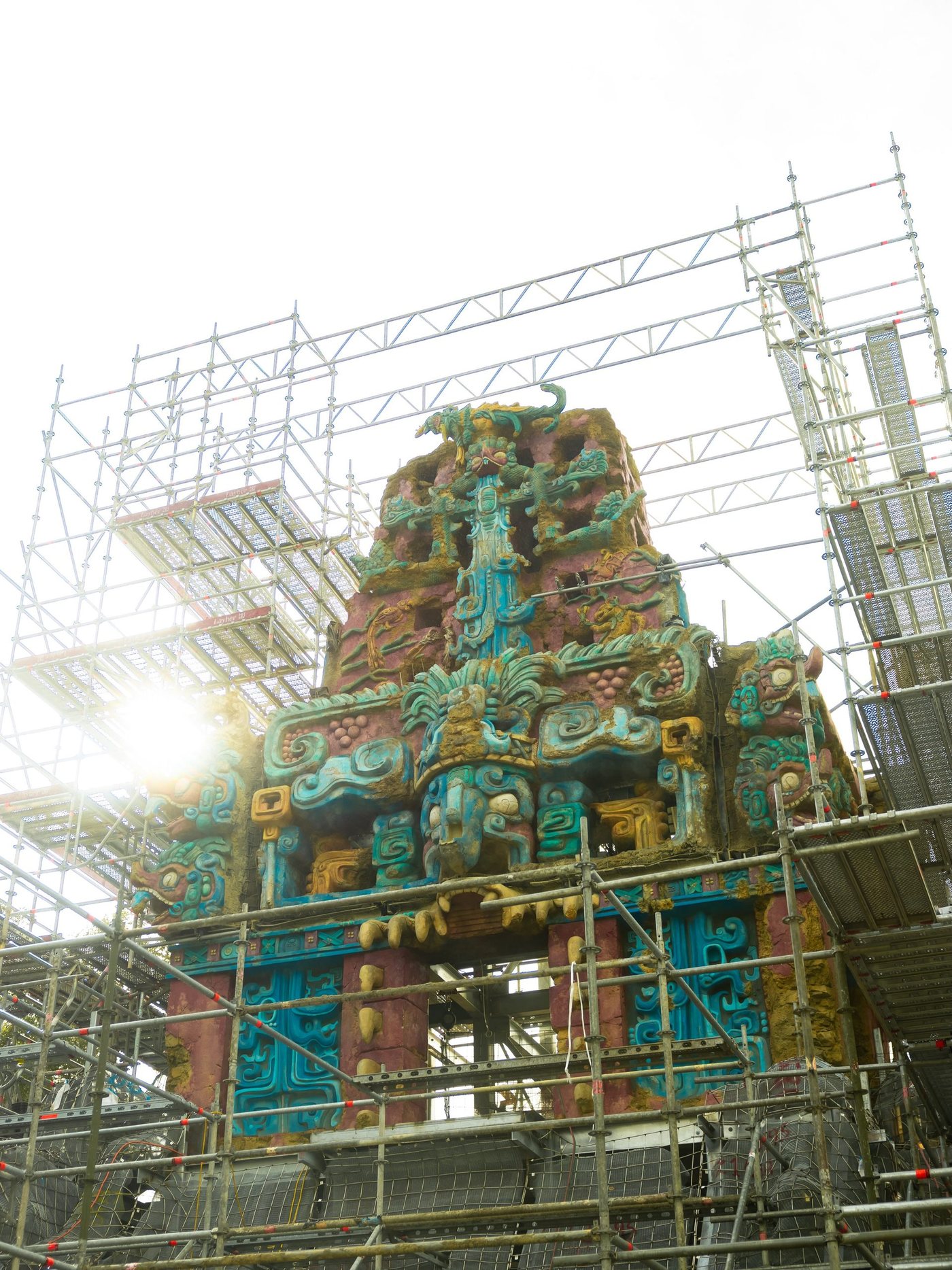 The upper tiers of a Maya-style temple covered in carved turquoise, red and gold rockwork, surrounded by metal scaffolding, with the sun flaring behind it