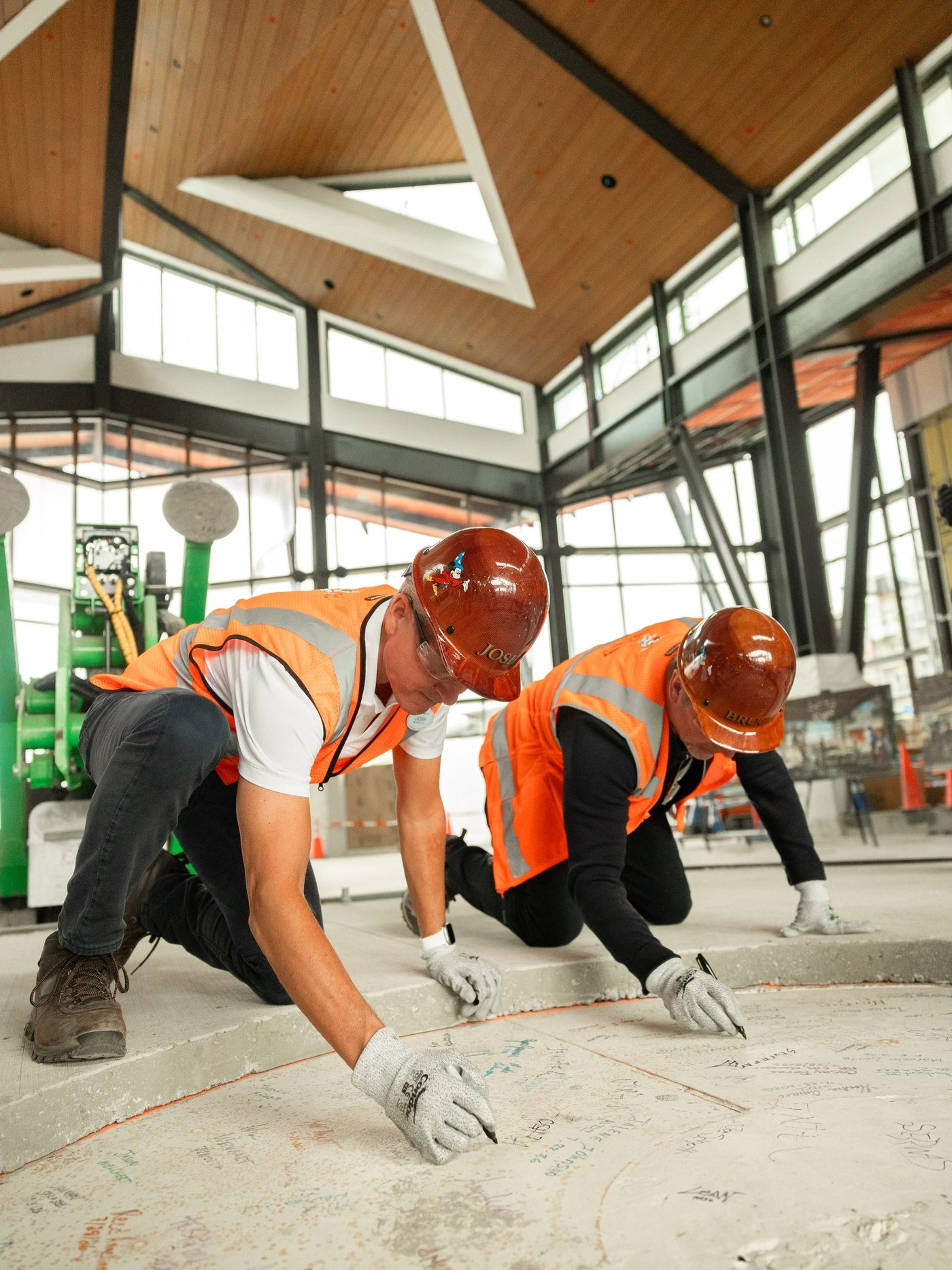 Two people in orange safety vests and brown hard hats kneel on a concrete floor covered in handwritten signatures, one hard hat labeled JOSH, inside a tall room with a wood-paneled ceiling, a triangular skylight and walls of windows