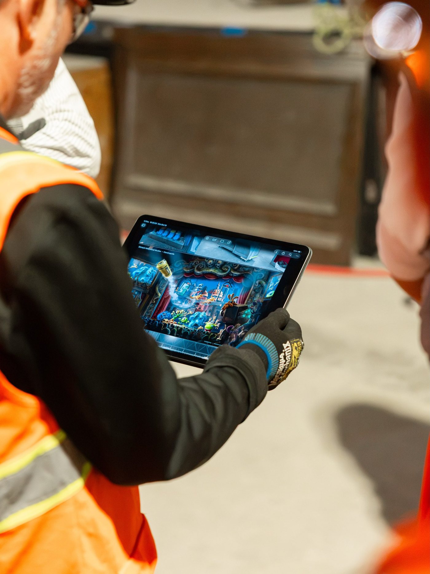 An Imagineer in an orange safety vest and work gloves holding a tablet that shows colorful concept art of a theater stage full of monsters in front of an audience