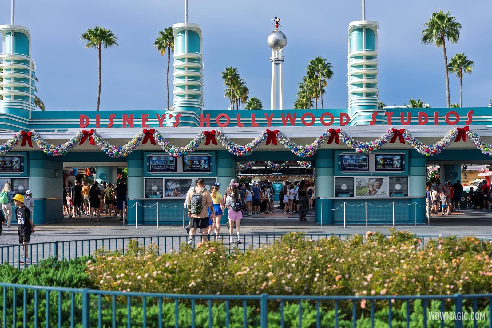 The teal art deco entrance of Disney's Hollywood Studios hung with silver garland, colorful ornaments and red bows, with guests walking toward the turnstiles and palm trees behind