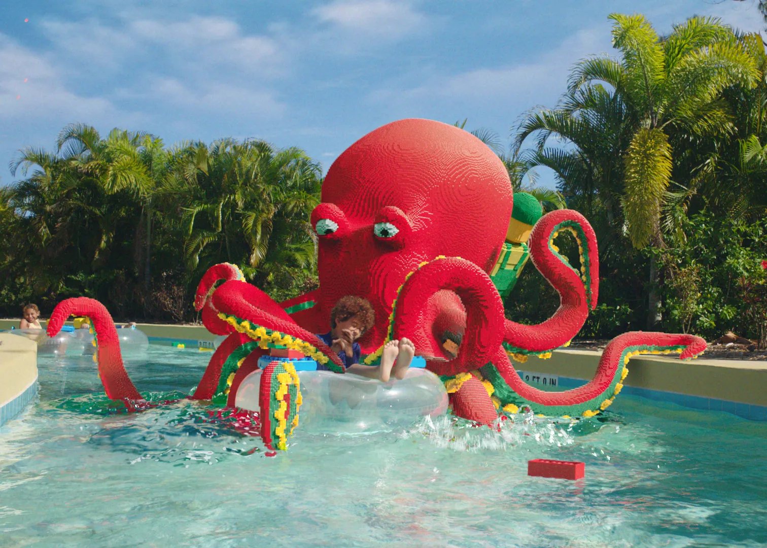 A giant red LEGO-style octopus sculpture with curling tentacles looms over a child riding a clear tube through the water park's river channel, with palm trees behind