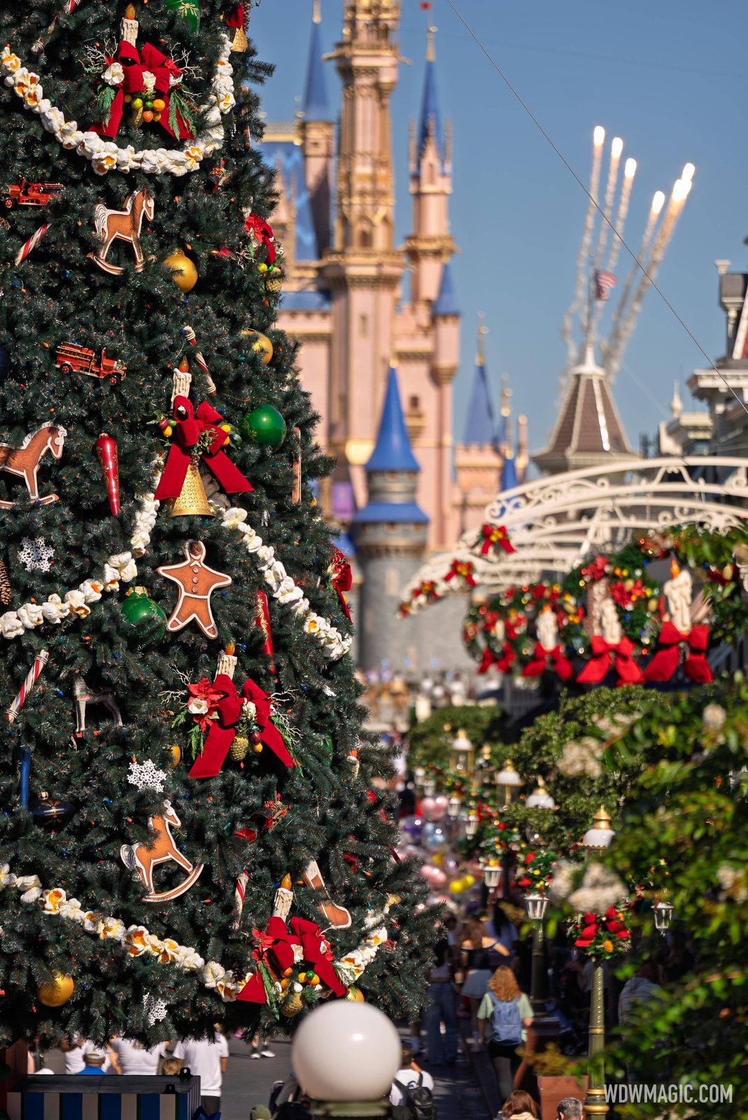 Close-up of a Christmas tree decorated with popcorn garlands, gingerbread men, rocking horses and red bows, with Cinderella Castle and wreath-hung Main Street buildings in the background under a blue sky