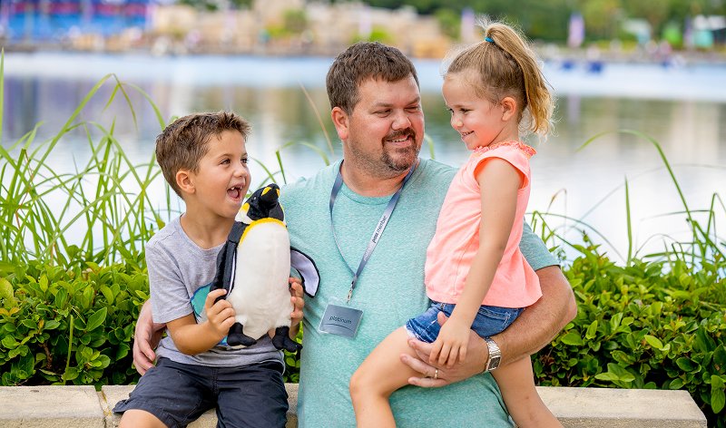 A smiling father wearing a lanyard pass holds his son, who has a penguin plush, and his daughter on his knee beside the lake at SeaWorld Orlando
