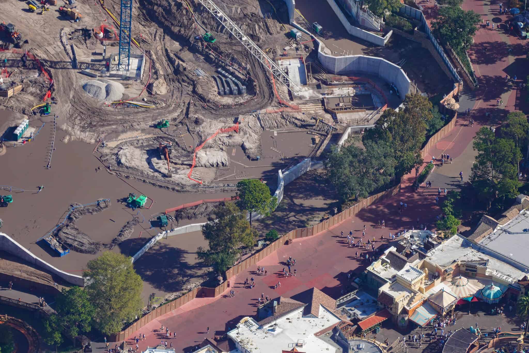 Aerial view of the muddy Piston Peak construction site with standing water, pumps and excavators, a curved concrete retaining wall with fresh concrete and formwork inside it beneath the white crane jib, and a guest walkway running along the construction walls at bottom right