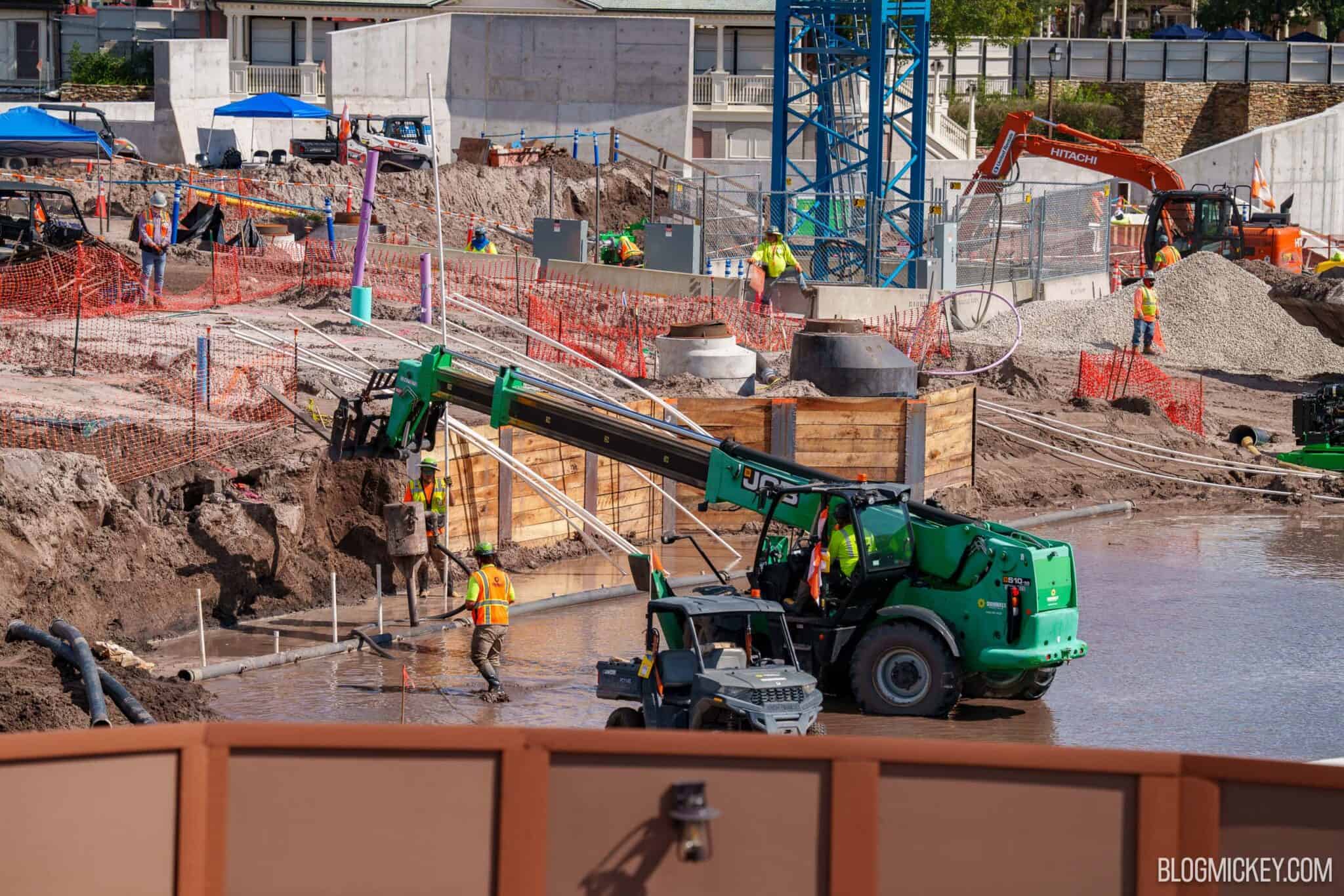 A green JCB telehandler holds a concrete bucket over an excavation as a worker in an orange vest guides the hose, with standing water around the machine, a timber-shored pit with two round vaults behind it, and the blue tower crane in the background
