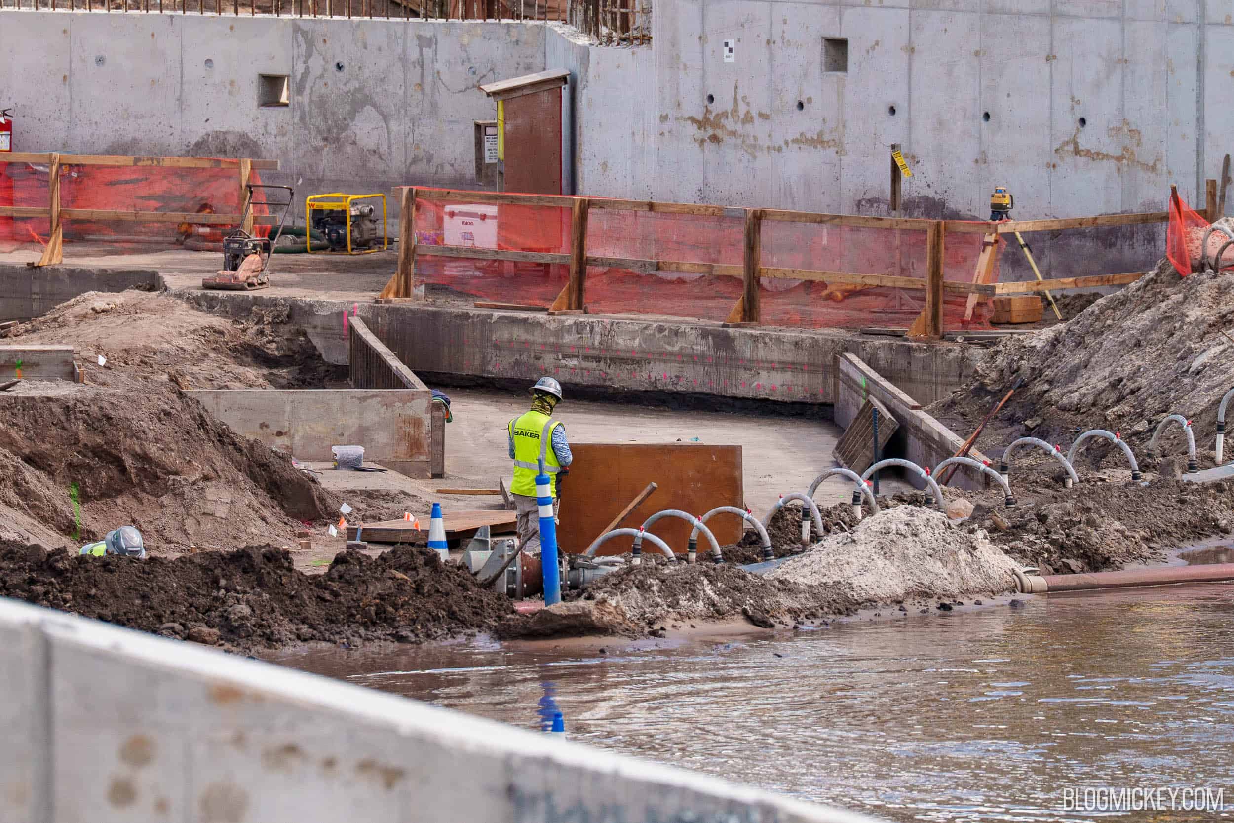 A worker in a hard hat and yellow BAKER safety vest stands on a new concrete slab ringed by low concrete walls and wooden forms, with a row of arched hoses along the muddy edge of standing water in the foreground and the tall weathered retaining wall behind