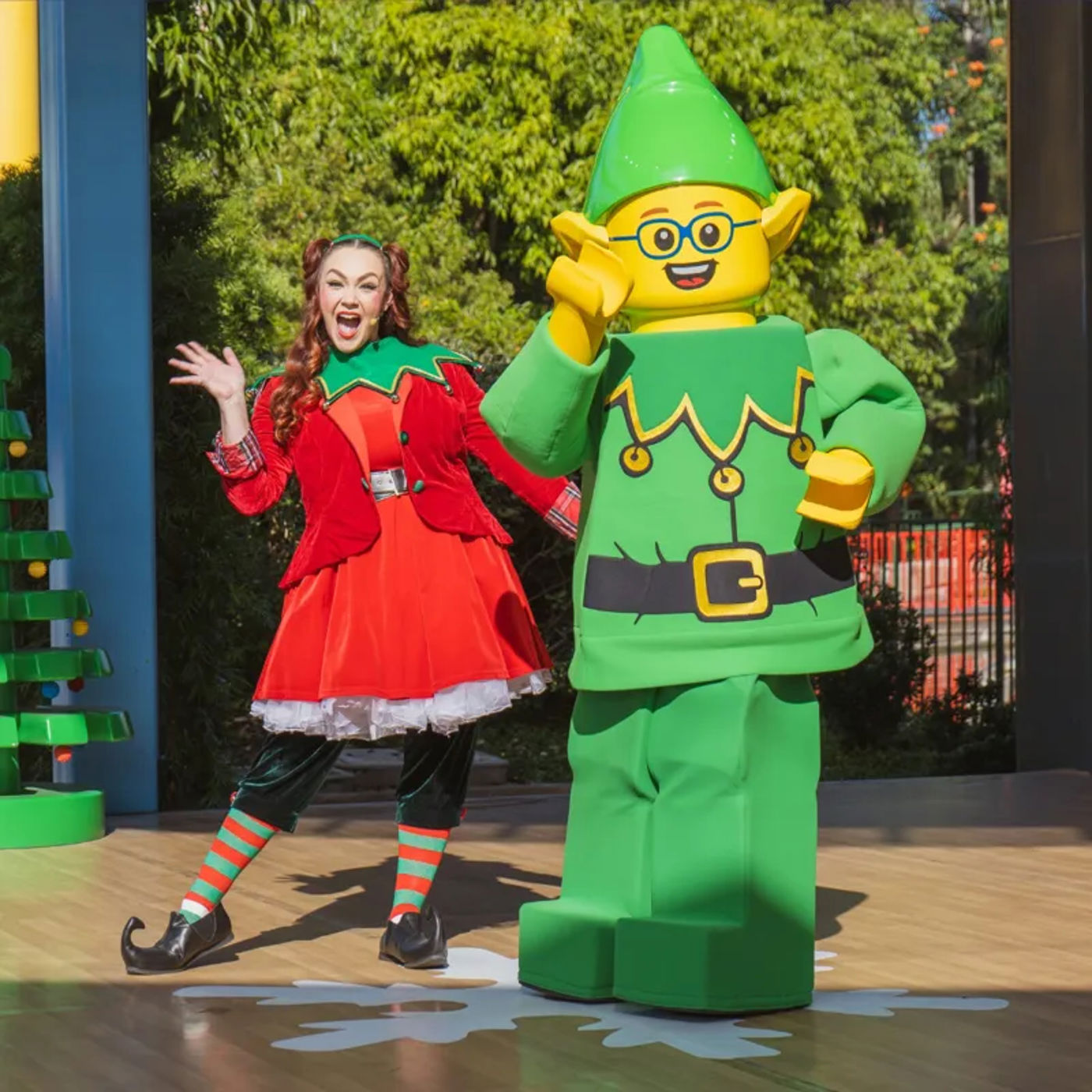 A performer in a red and green elf costume with striped stockings waves from an outdoor stage beside a costumed LEGO elf character in green with glasses and a pointed hat, with a LEGO Christmas tree at left