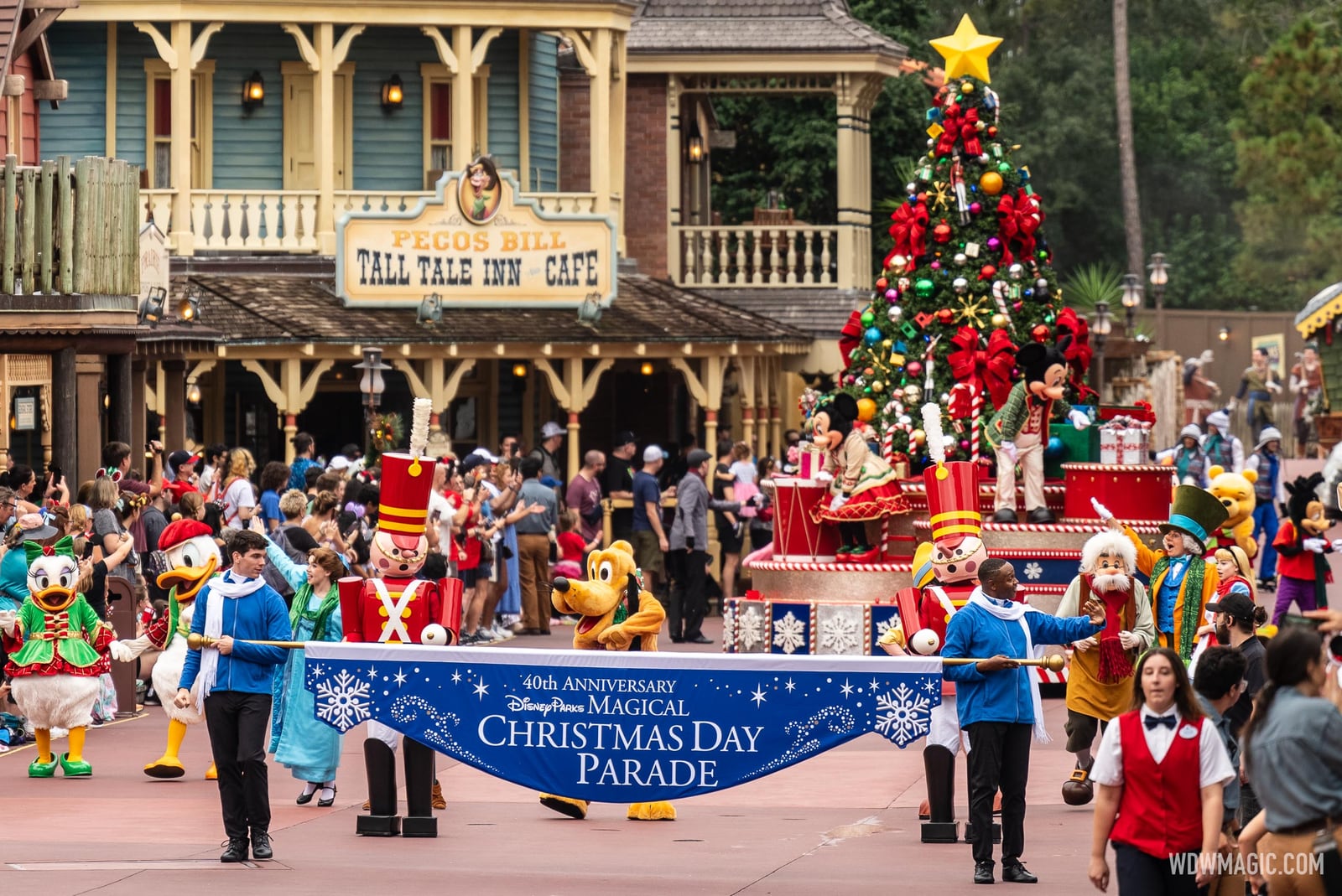 Performers in blue sweaters and white scarves carry a long blue banner reading 40th Anniversary Disney Parks Magical Christmas Day Parade through Frontierland in daylight, with Donald Duck, toy soldiers, Pluto and a Christmas tree float behind them and the Pecos Bill Tall Tale Inn and Cafe in the background