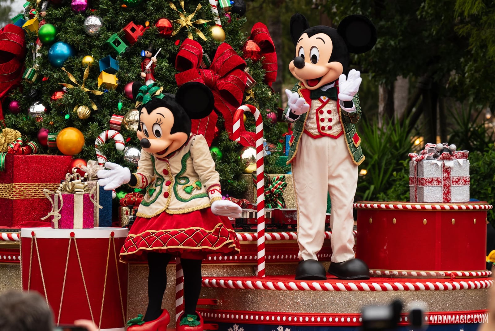 Minnie Mouse in a cream jacket with green trim and Mickey Mouse in a green jacket over a cream vest wave from a float decorated with a large Christmas tree, red bows, drums and wrapped presents