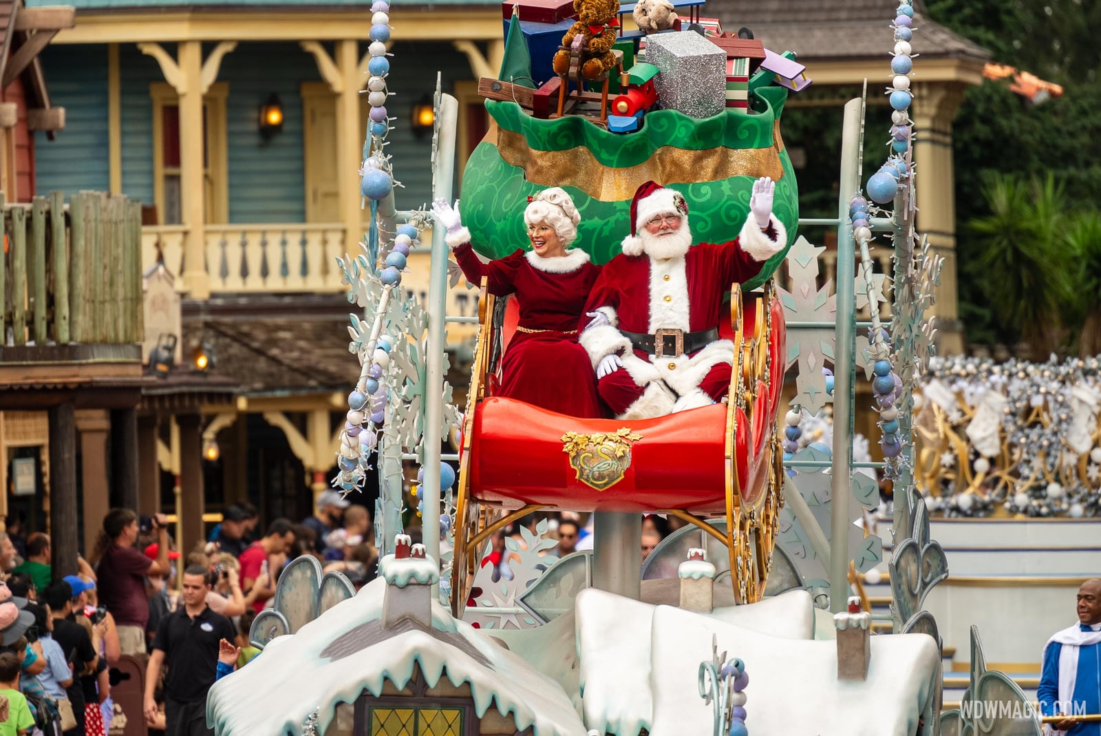 Santa Claus and Mrs. Claus wave from a red sleigh atop a snowy float as it passes Western-style buildings in Frontierland, with guests lining the route