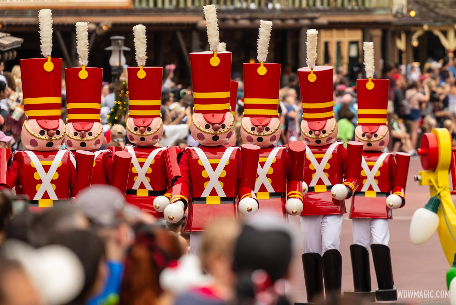 A line of large toy soldier characters with tall red hats, rosy cheeks and white crossed sashes march past a crowd of guests in daylight