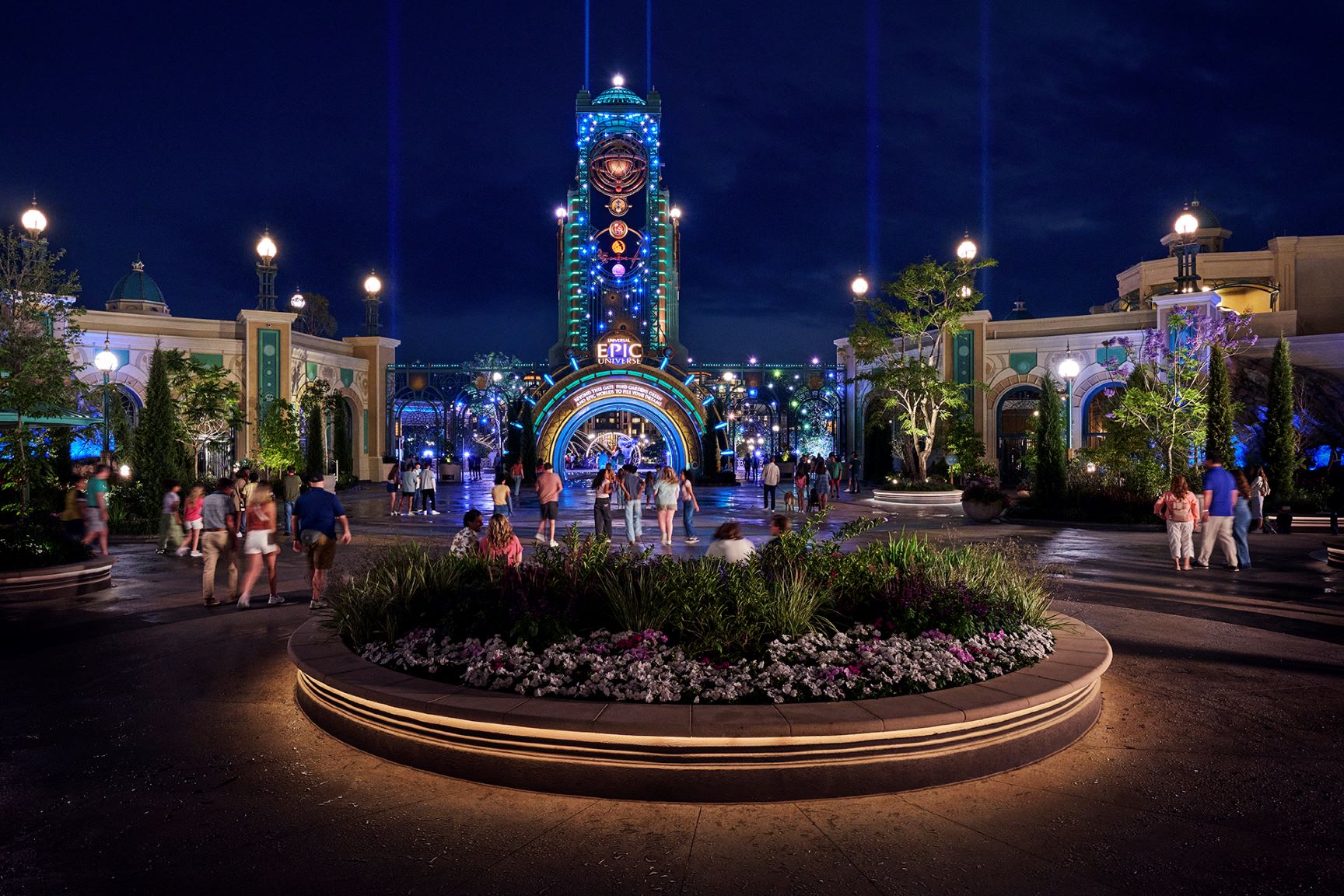 The Epic Universe entrance at night, with the illuminated EPIC entrance tower and portal arch glowing blue and green beyond a lit circular planter and guests walking on the plaza